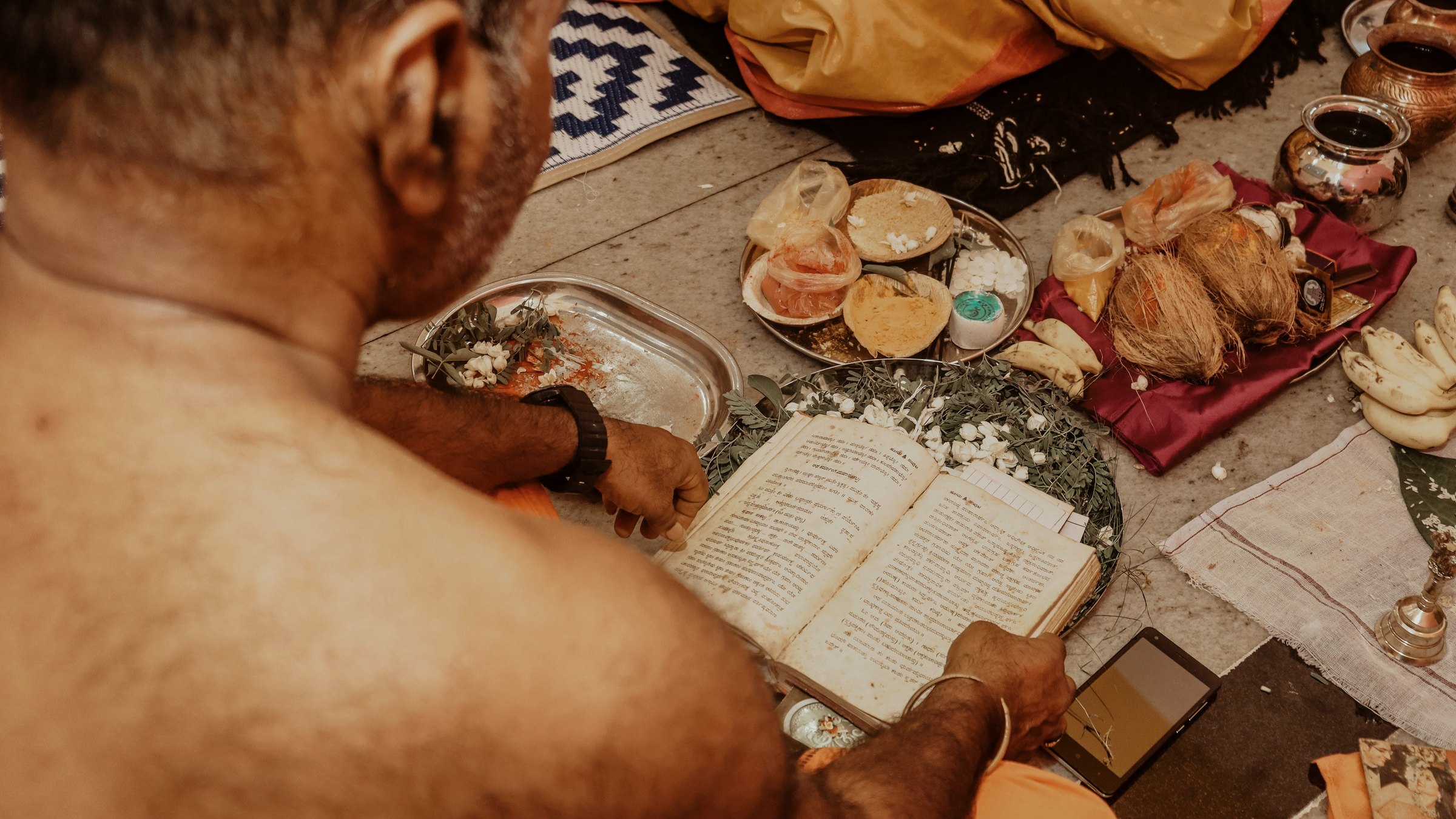 An elderly man reading from a text beside plates of offerings